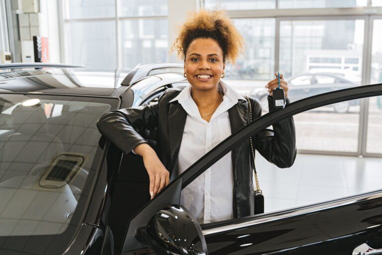 Smiling woman with car keys standing beside her new vehicle in a dealership showroom.
